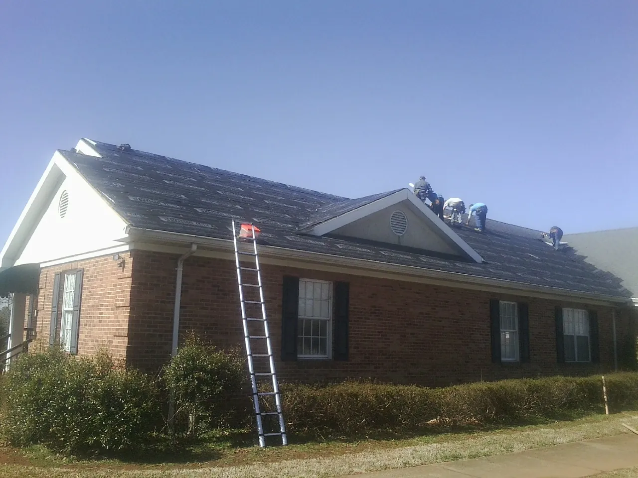 Crew tearing shingles off a brick ranch roof with a ladder set — Metal Roof Repair work by our crew in Arco